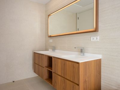 Modern floating wooden bathroom vanity with dual integrated sinks and chrome faucets. Features a backlit mirror with a wooden frame and textured wall tiles.