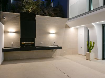 Modern integrated outdoor kitchen with BBQ, stone countertops, and dining area, framed by native vegetation and panoramic Mediterranean sea views.