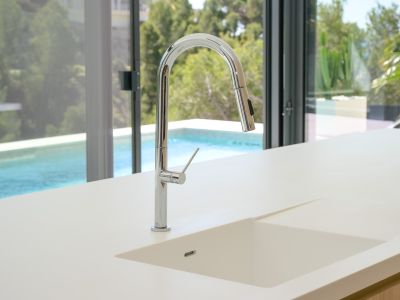 Modern kitchen featuring a light countertop with an integrated sink and a sleek chrome pull-out faucet. Large glass doors offer views of the swimming pool and lush greenery.
