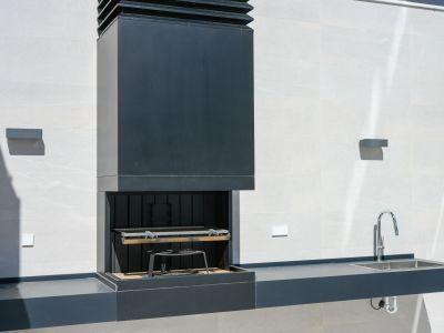 Modern outdoor kitchen featuring a dark integrated barbecue with a sleek hood, a matching counter with a stainless steel sink, and a chrome faucet, set against a light tiled wall.