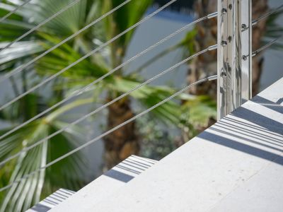 Modern outdoor staircase detail: light-toned steps, polished metal railing with steel cables. Sunlight casts sharp parallel shadows, framed by lush palm foliage.