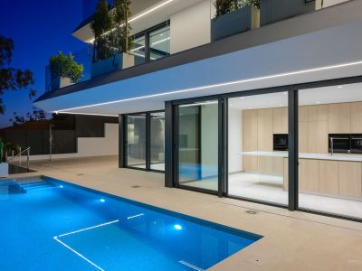 Night view of the modern villa, showcasing the illuminated blue pool and sleek architecture. A light wood outdoor kitchen is visible through the large glass doors.