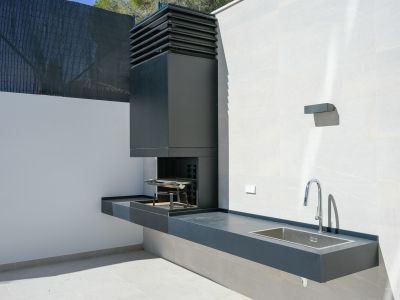 Outdoor kitchen featuring an integrated dark grey BBQ unit, a sleek countertop with a stainless steel sink, and a modern chrome faucet, set against light tiled walls.
