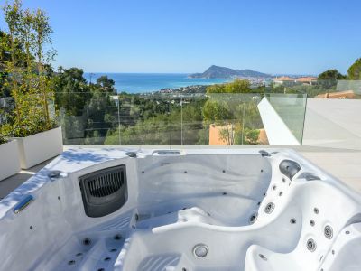Outdoor spa on a tiled terrace offers panoramic views of the Mediterranean Sea, coastal town, and distant mountains. Glass railings ensure an unobstructed vista, complemented by lush greenery.