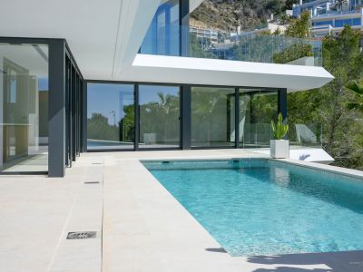 Private swimming pool with blue mosaic tiles and light stone terrace, bordered by large glass doors connecting to the villa's modern interior.