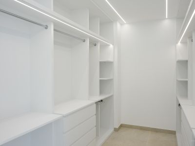 Spacious walk-in closet featuring custom white cabinetry, chrome hanging rails, and integrated LED strip lighting for optimal illumination. Includes multiple shelves and drawers, set on light-toned tiled flooring.