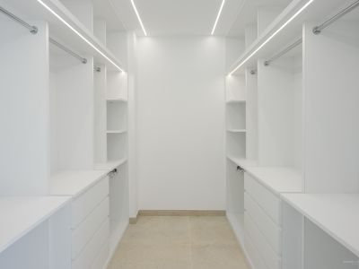 Spacious white walk-in closet featuring custom shelving, drawers, and hanging rails, illuminated by integrated LED strips and light tiled flooring.