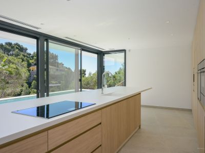 The modern kitchen features a large island with a white countertop, integrated induction hob, and chrome faucet. Sliding glass doors offer views of lush Mediterranean greenery and a private pool.