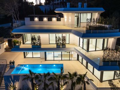 The modern villa's multi-level architecture is illuminated at dusk, showcasing its vibrant blue pool and expansive terraces with glass railings.