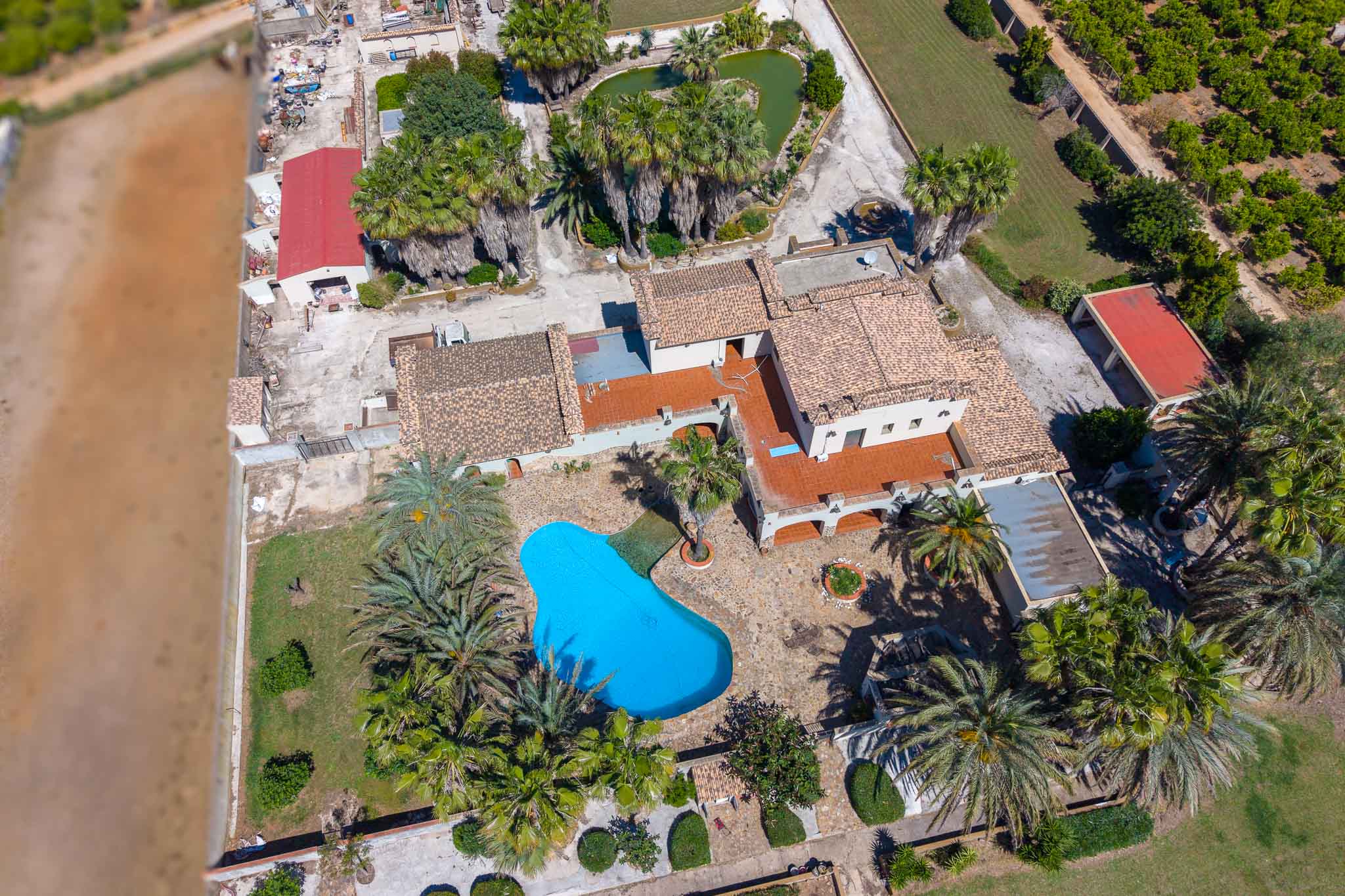 Aerial view of the estate featuring an irregular-shaped blue swimming pool, surrounded by palm trees and gardens. Multiple buildings with terracotta roofs and a heart-shaped pond are visible.