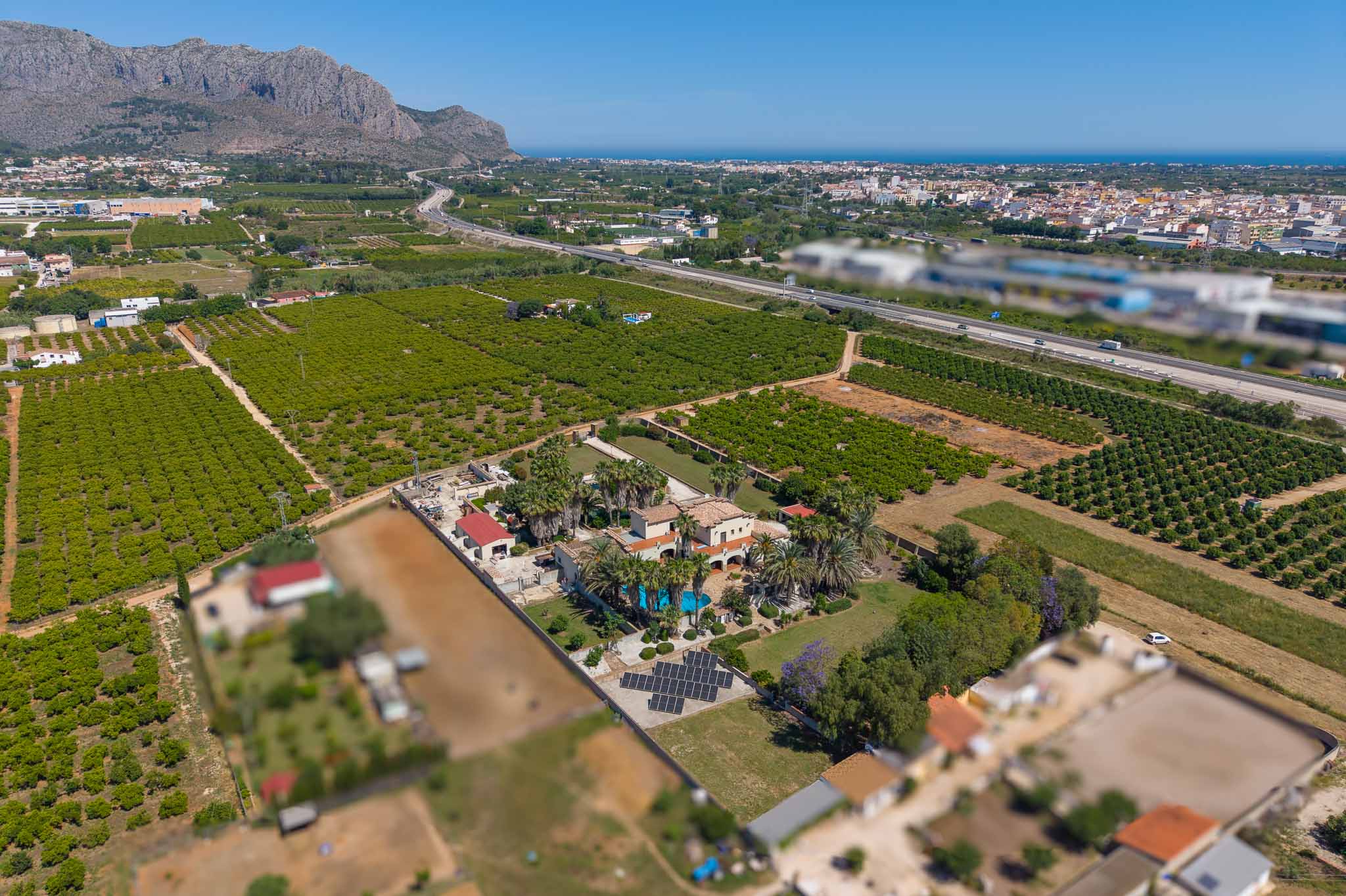 Aerial view of the estate featuring the main house, swimming pool, extensive gardens, and a large solar panel array, set amidst orange groves with distant sea and mountain views.