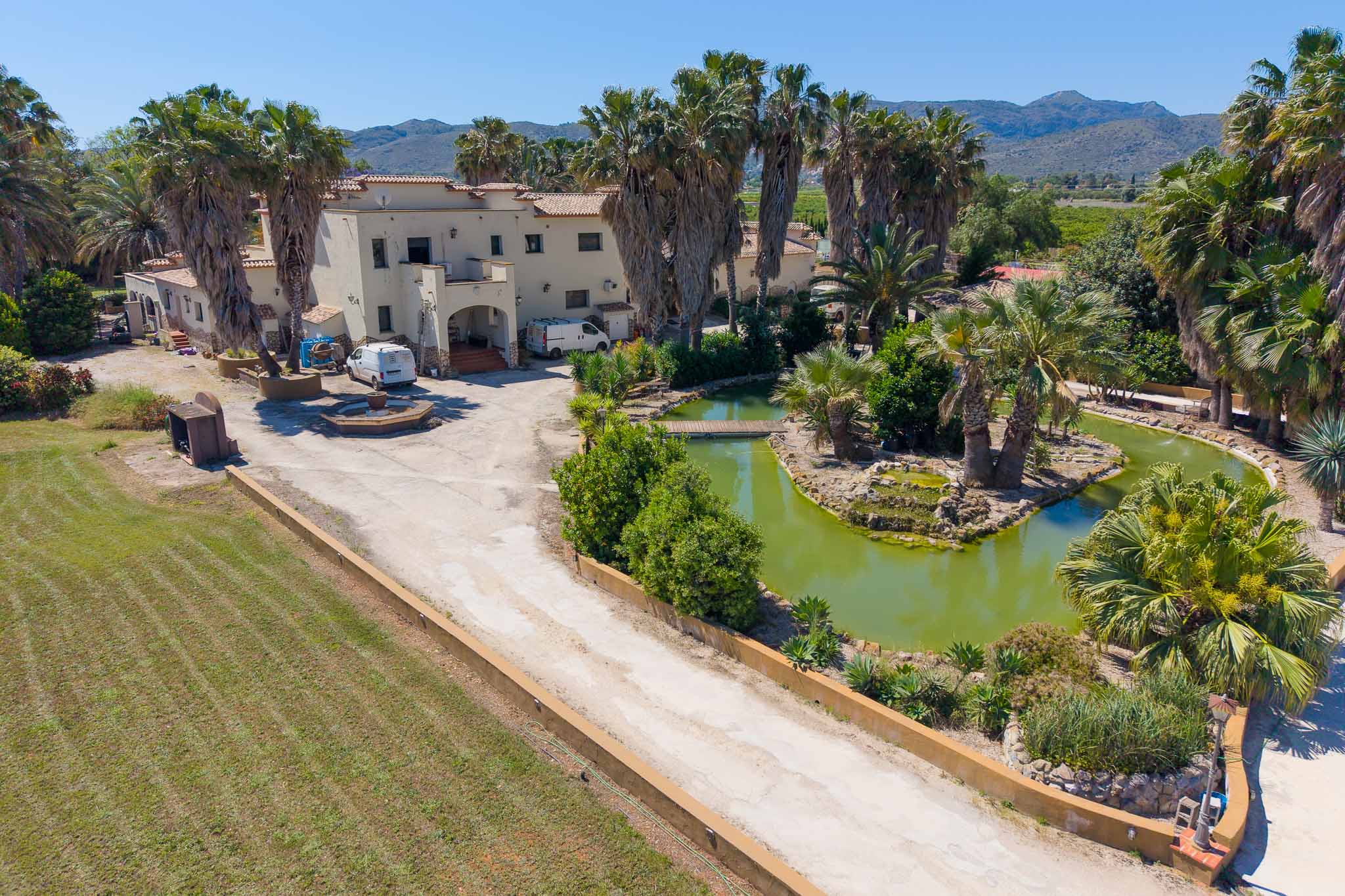 Aerial view of the Mediterranean-style main villa, surrounded by extensive palm trees, a pond with a bridge, and a long driveway, set against a mountain backdrop.