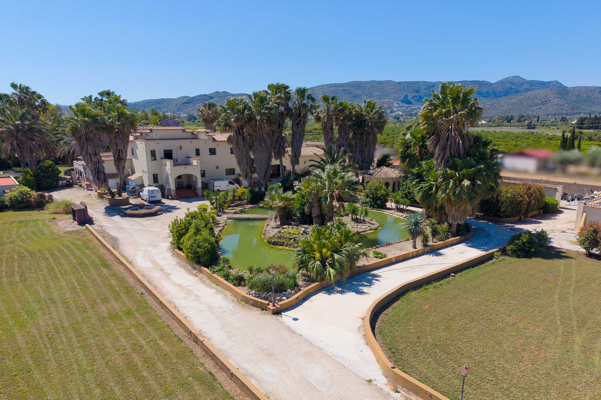 Aerial view showcasing the extensive property with a winding driveway, a large central pond with a bridge, lush palm trees, and the main villa against a mountain backdrop.