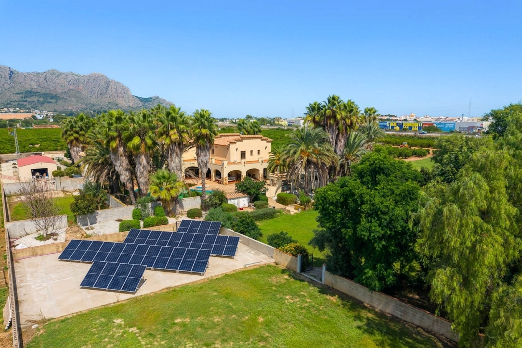 Aerial view showcasing the traditional villa surrounded by palm trees and lush gardens, featuring a prominent solar panel installation and distant mountain vistas under a clear sky.