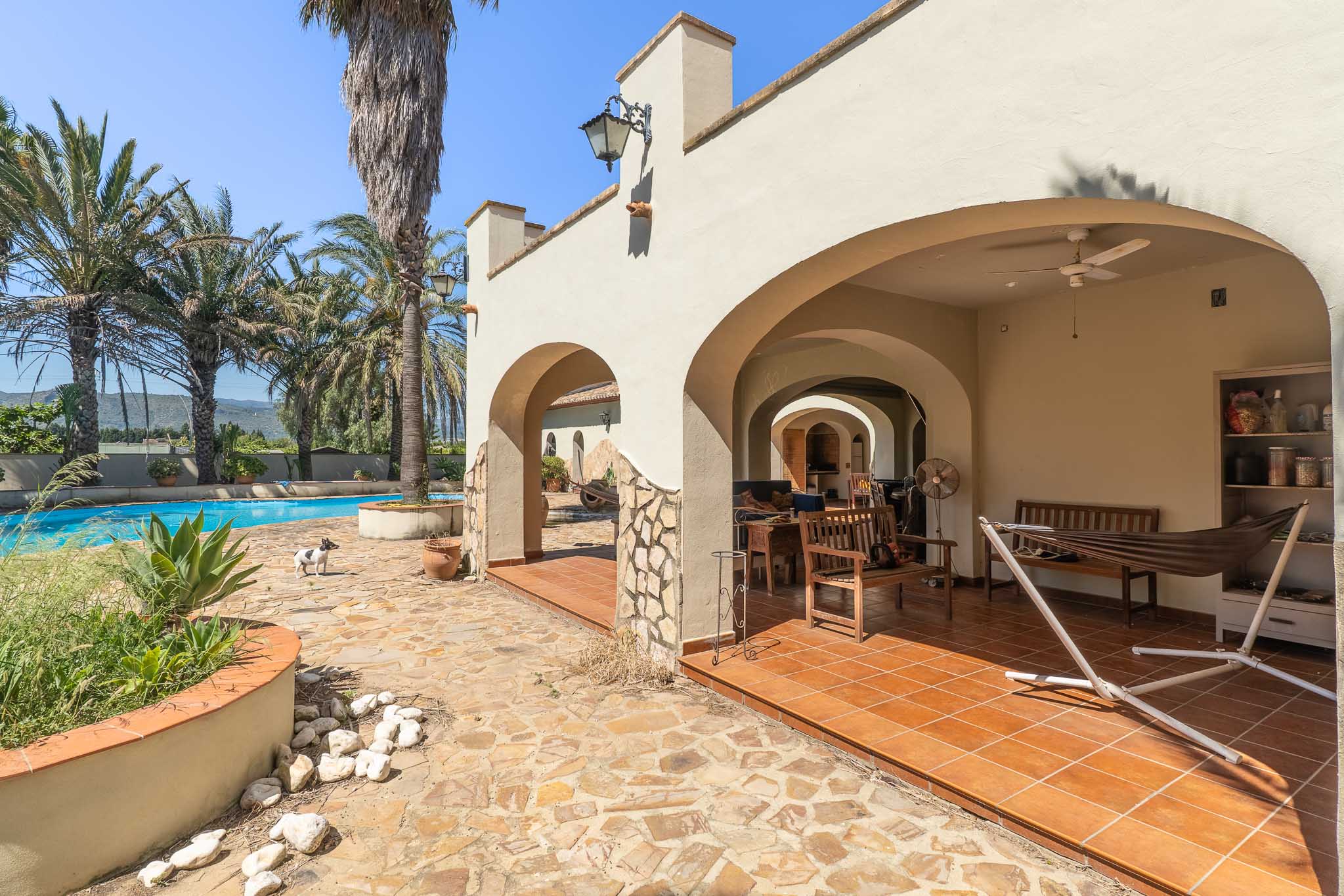 Arched covered terrace with terracotta tiled floor, featuring outdoor furniture and a hammock, adjacent to the swimming pool and palm trees with mountain views.