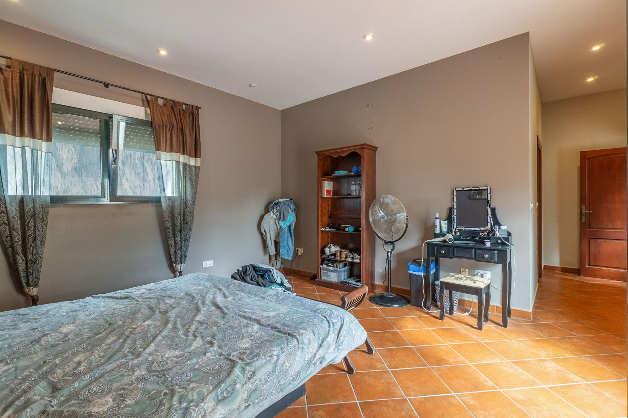 Bedroom featuring a bed, windows with brown and sheer curtains, and terracotta tiled flooring. Includes a black dressing table, wooden bookshelf, and standing fan.