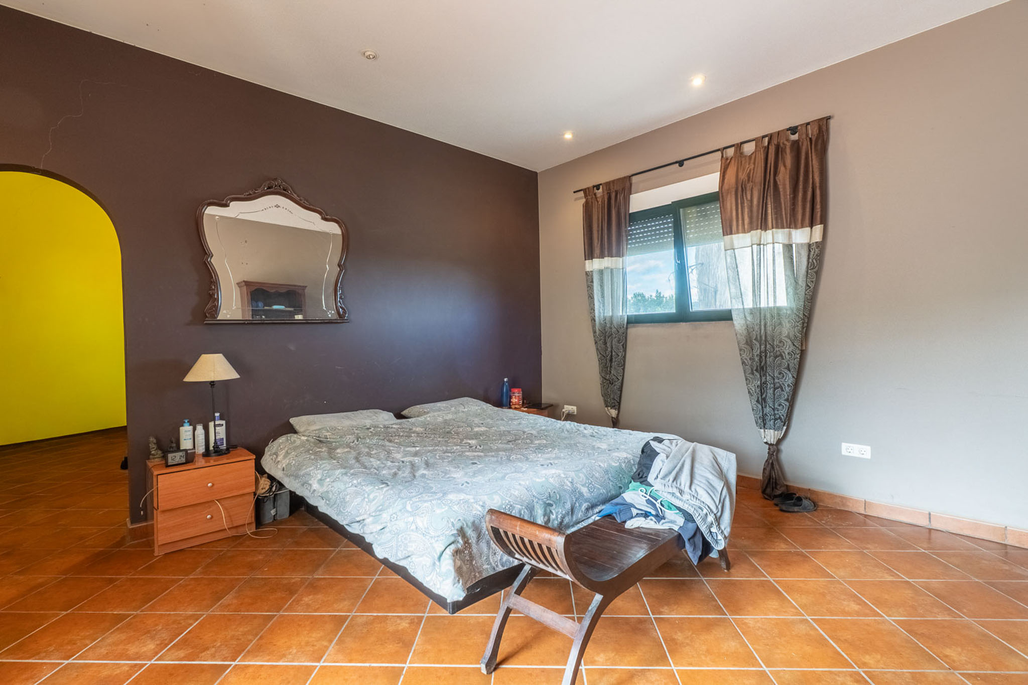 Bedroom featuring a double bed, terracotta tiled floor, and two-tone walls. A window with blinds and curtains provides natural light, complemented by a decorative mirror.