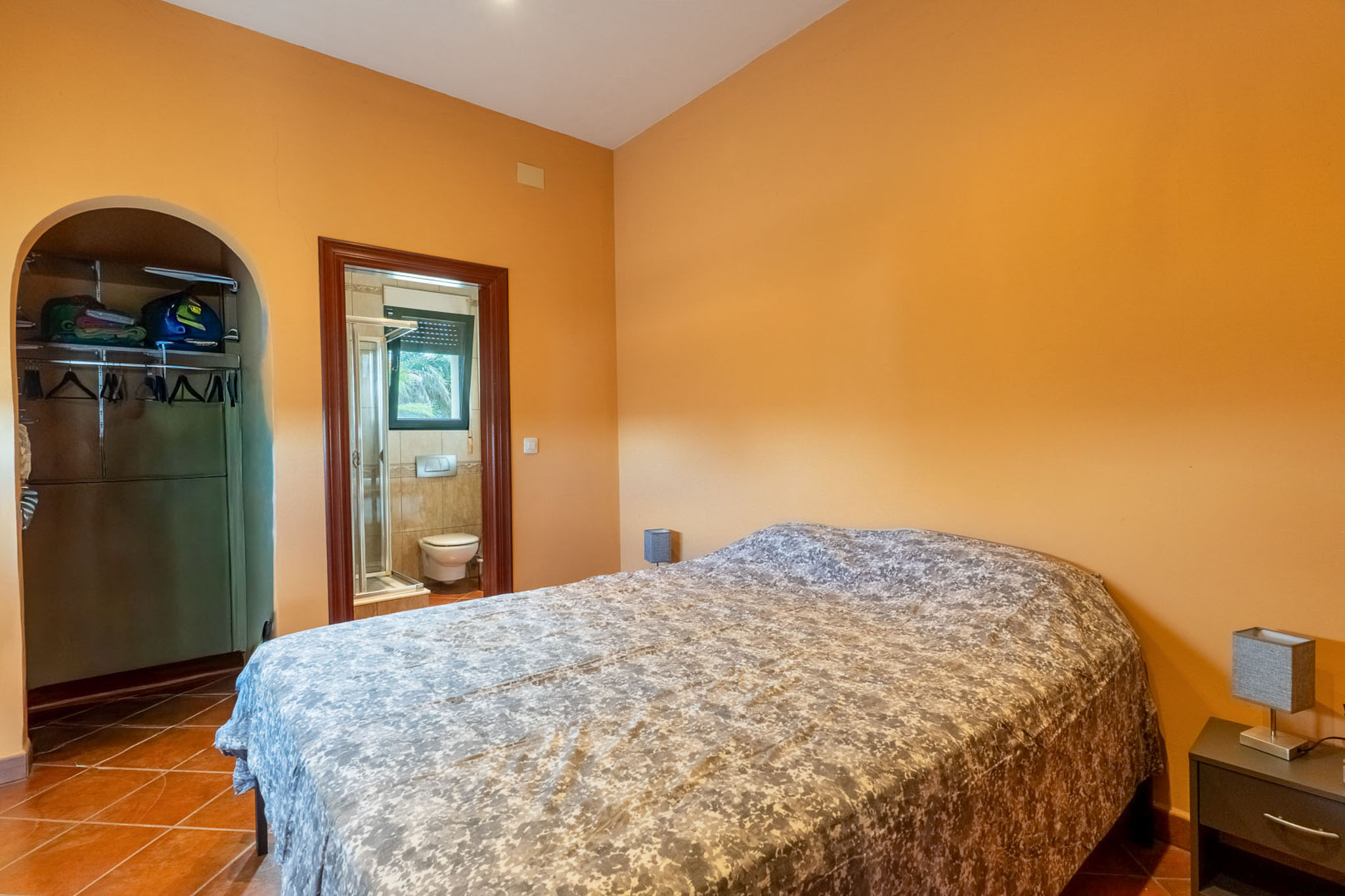 Bedroom featuring warm orange walls and terracotta tiled flooring, furnished with a double bed. An arched opening leads to an open closet, with an en-suite bathroom visible through the door.