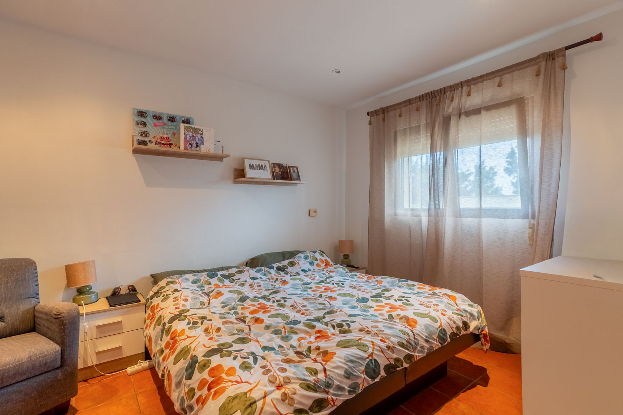 Bright bedroom with a double bed, colorful leaf-patterned duvet, and terracotta tiled floor. Natural light enters through a window with sheer curtains, complemented by a bedside table and wall shelves.