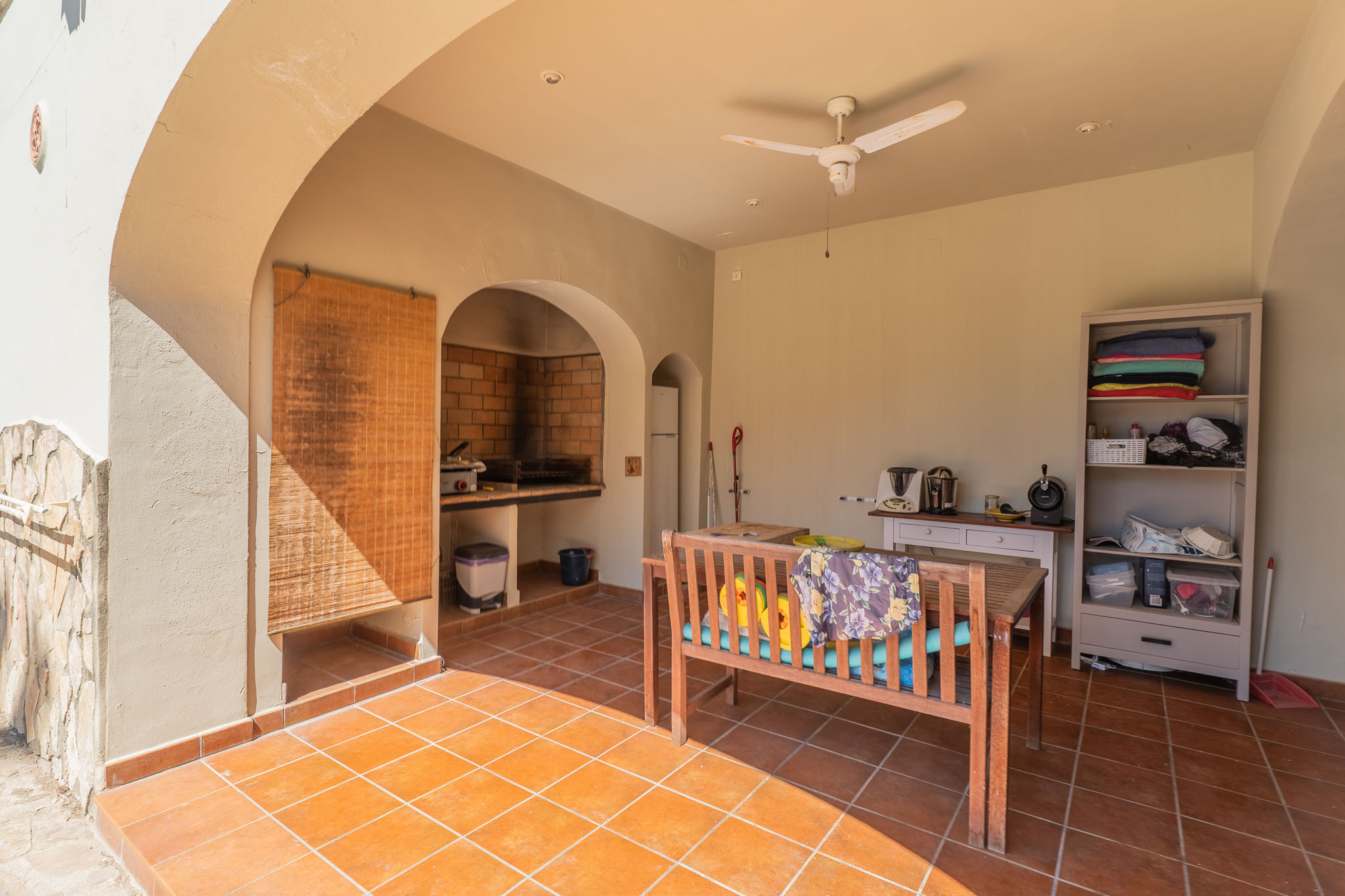Covered terrace featuring terracotta tiled flooring and a built-in brick barbecue. Includes a wooden dining bench, ceiling fan, and storage, perfect for outdoor entertaining.