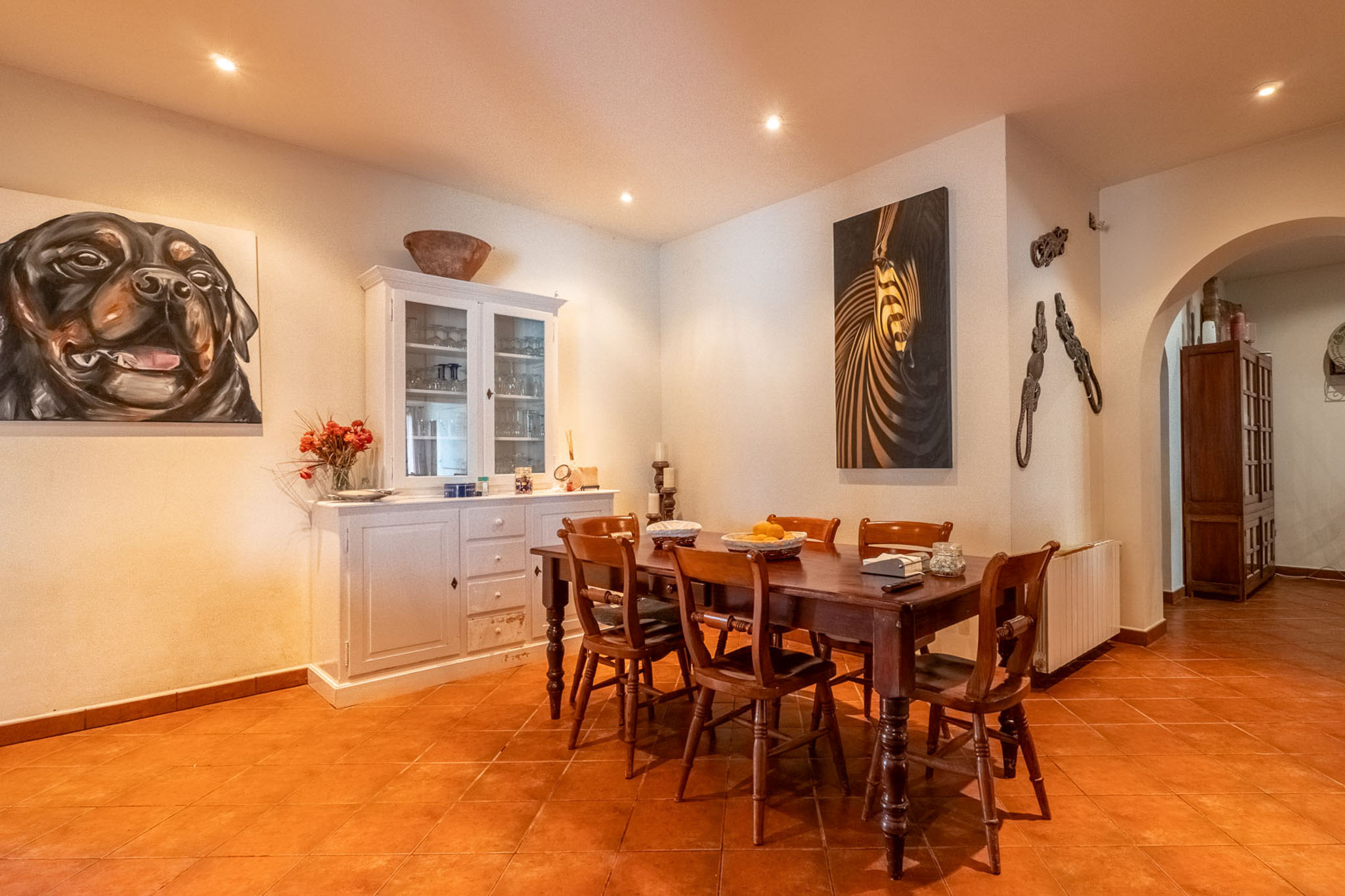 Dining room featuring a dark wooden table with six chairs, terracotta tiled floor, and white walls with recessed lighting. A white hutch and two large paintings adorn the space.