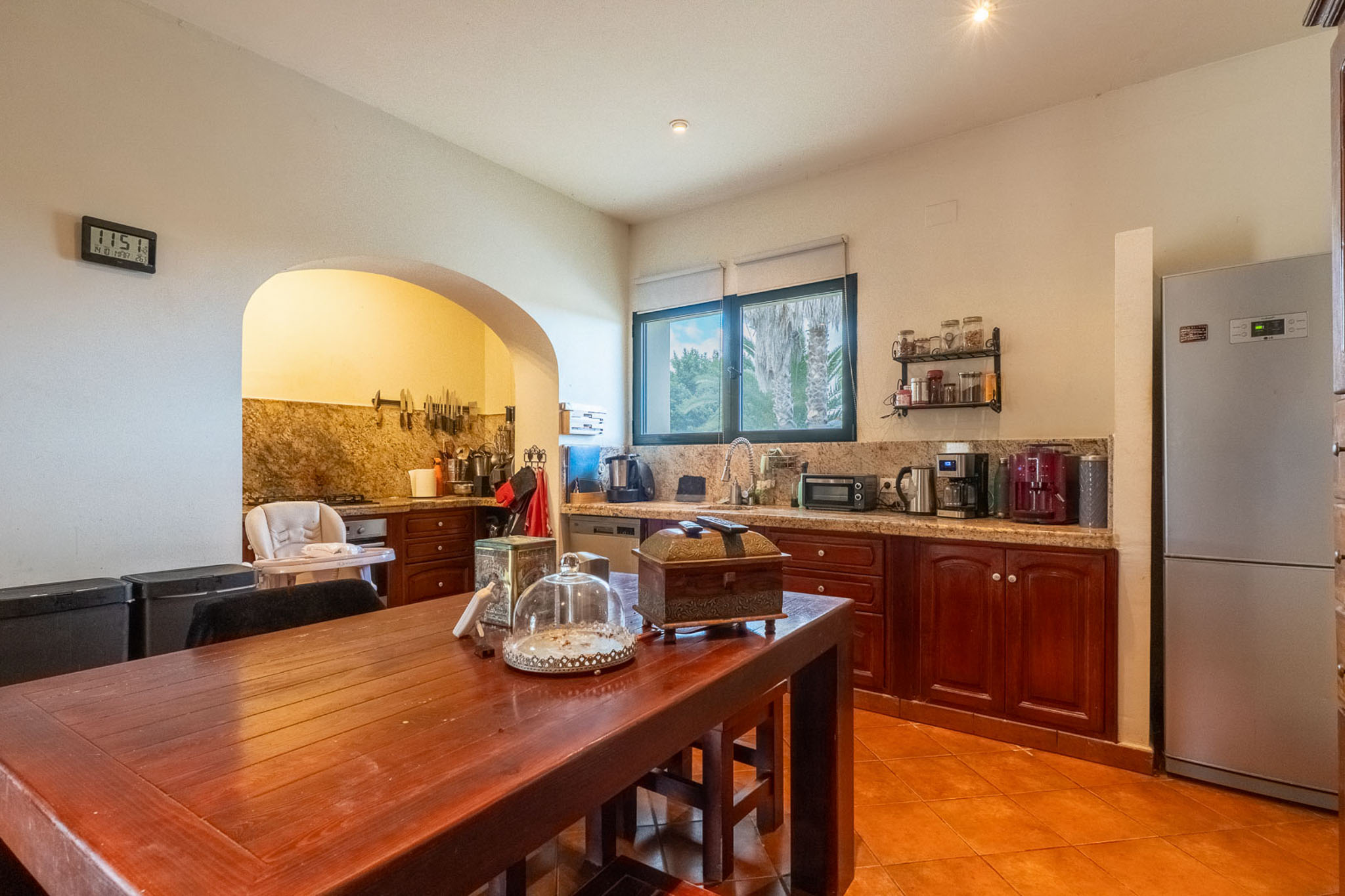 Equipped kitchen featuring granite countertops, dark wood cabinetry, terracotta floor tiles, and a central wooden dining table. An archway connects to an additional preparation area.