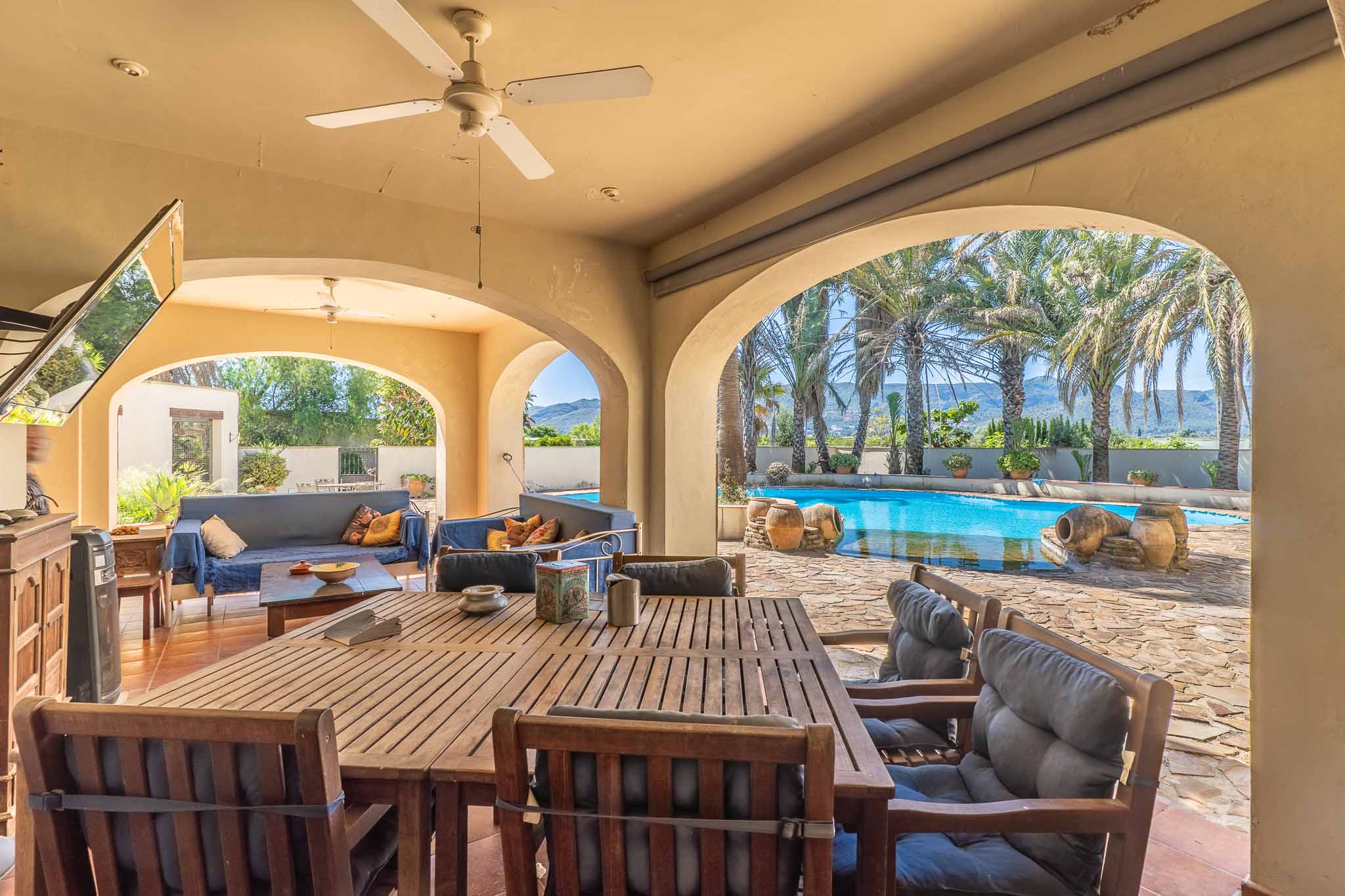 Expansive covered terrace with arches, featuring a wooden dining set and lounge area, offering direct views of the pool, palm trees, and distant mountains.