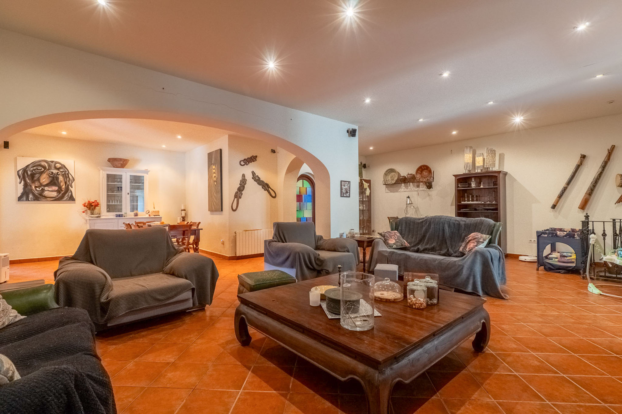 Expansive living room featuring terracotta tiled flooring and recessed ceiling spotlights. Includes a large sofa, armchairs, and a carved wooden coffee table, connecting to a dining area via archways.