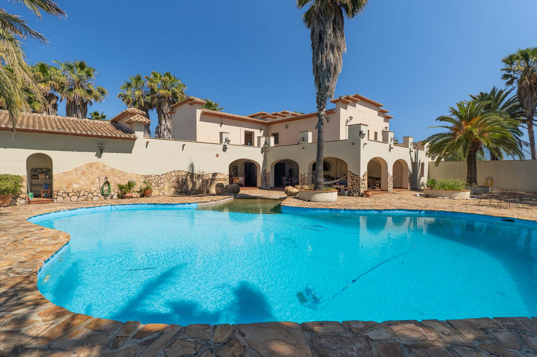 Exterior view of the Mediterranean villa featuring an irregular-shaped private pool with natural stone coping, surrounded by palm trees under a clear blue sky.