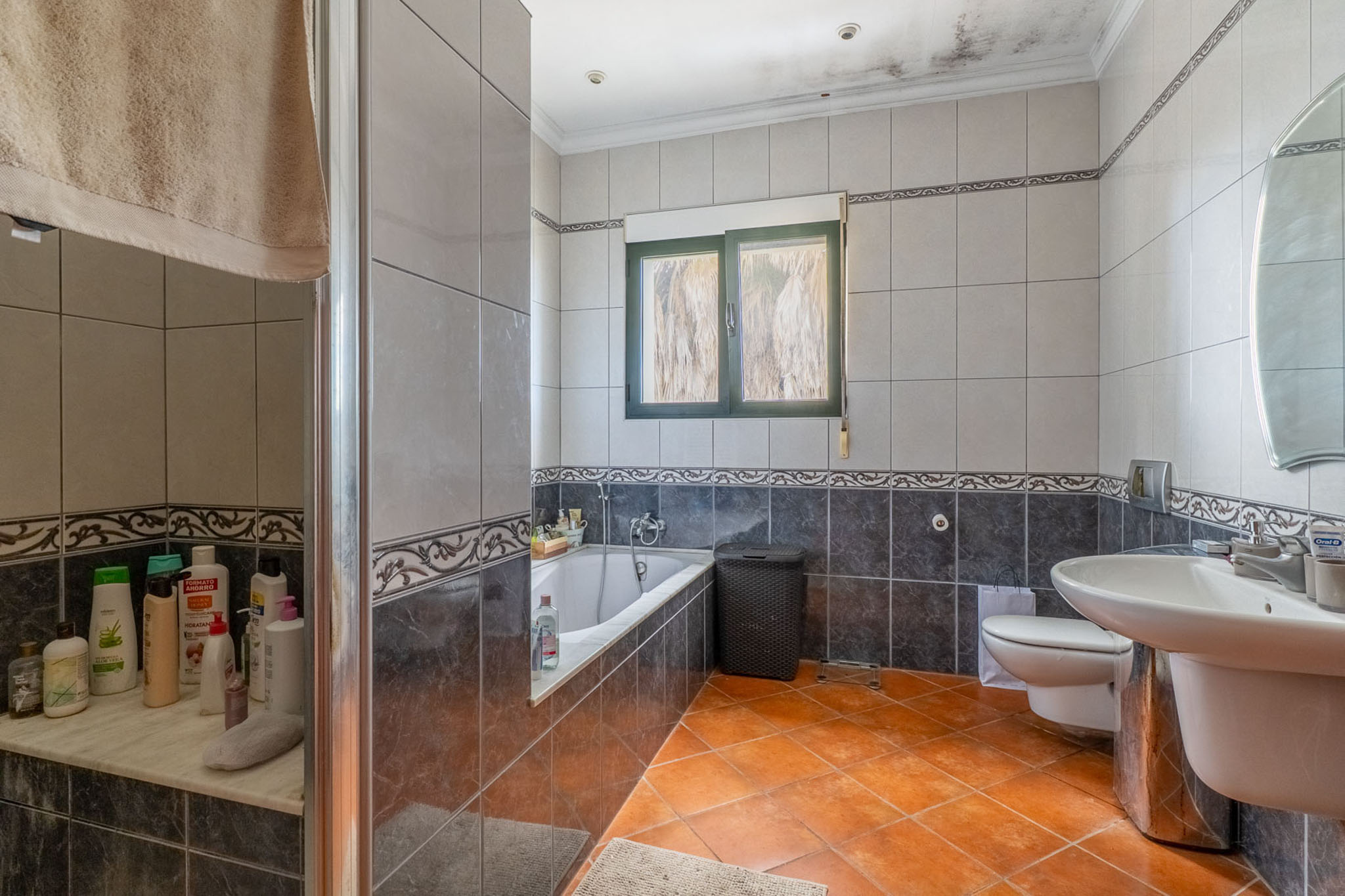 Full bathroom featuring terracotta floor tiles, grey wall tiles with decorative borders, a bathtub, glass-enclosed shower, sink, and a green-framed window.