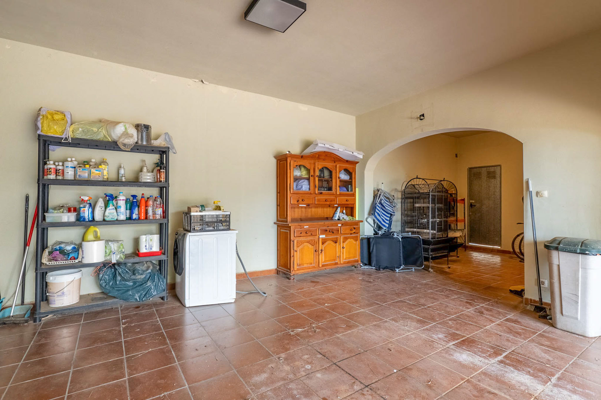 Functional basement utility room with terracotta tiled floor, a washing machine, and extensive shelving for storage, leading to an additional area through an archway.