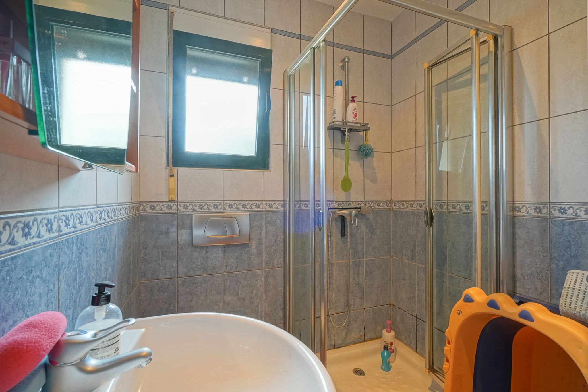 Functional bathroom featuring a ceramic sink, a glass-enclosed shower, and walls tiled in blue and beige. Natural light enters through the window.