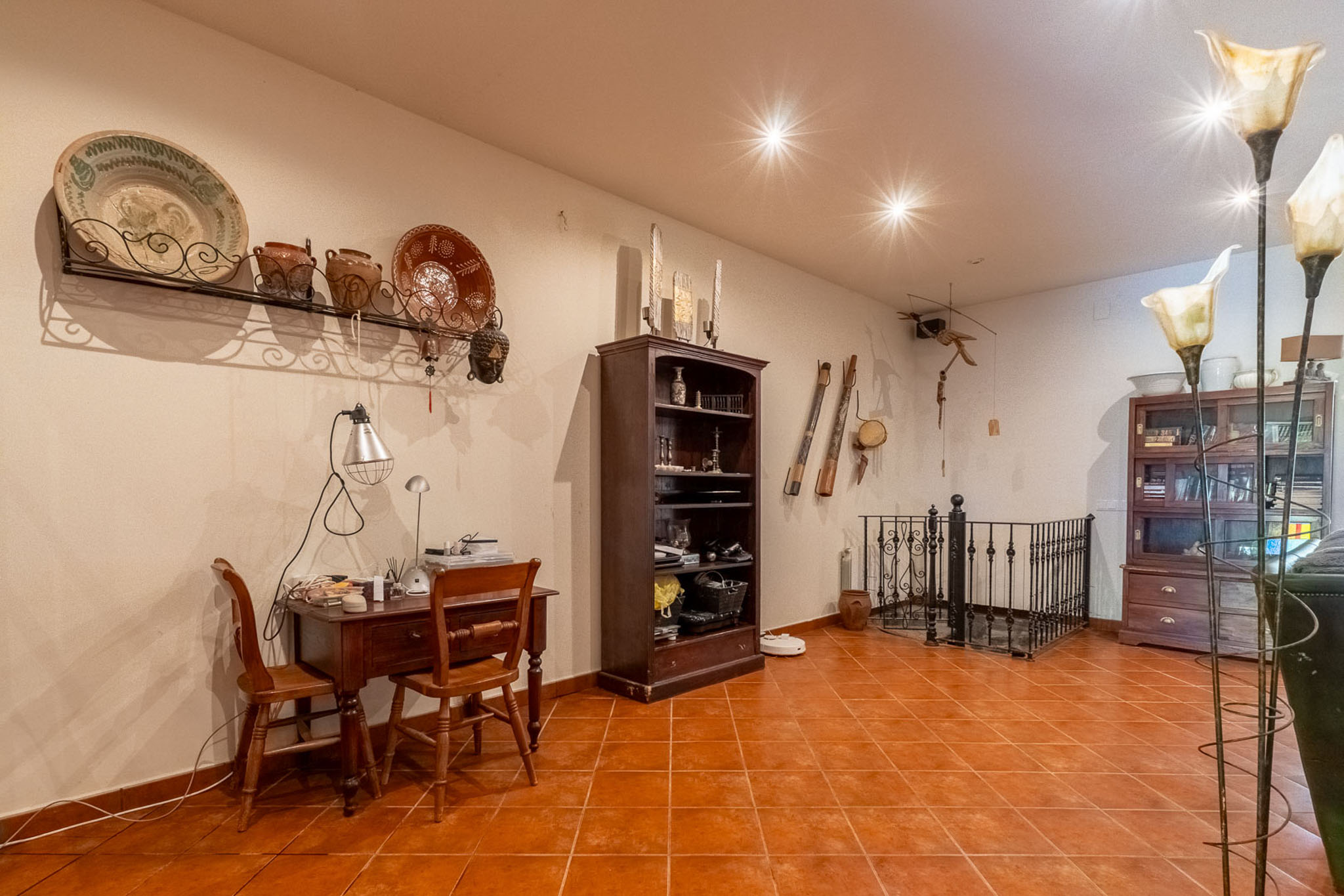 Functional home office area featuring a wooden desk, terracotta floor tiles, and traditional wall decor. The space includes a bookshelf and an elegant wrought-iron balustrade.