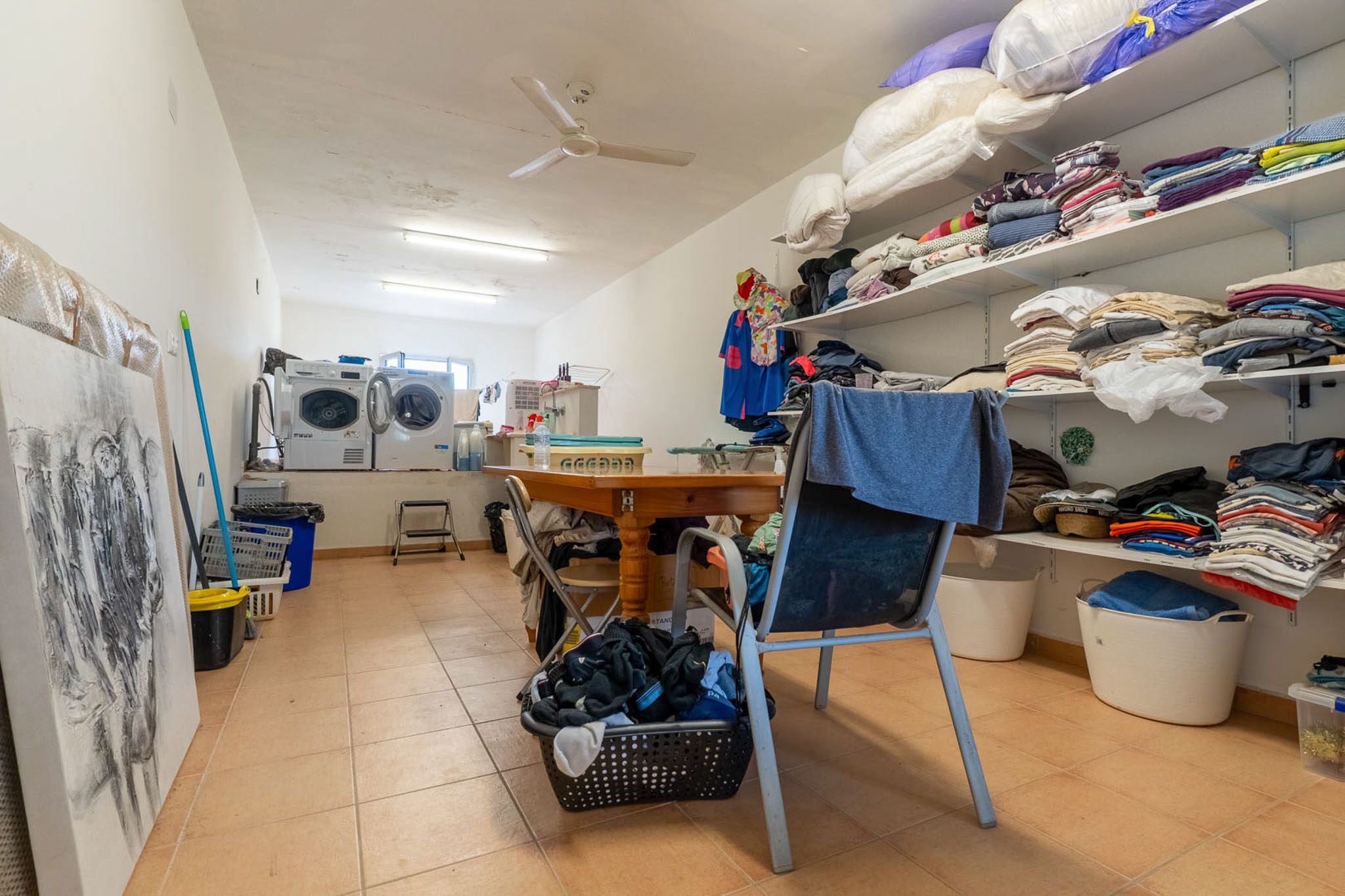 Functional utility room featuring two washing machines, a dryer, and extensive open shelving for laundry and storage. A central wooden table provides a practical workspace, complemented by tiled flooring and ceiling fan.