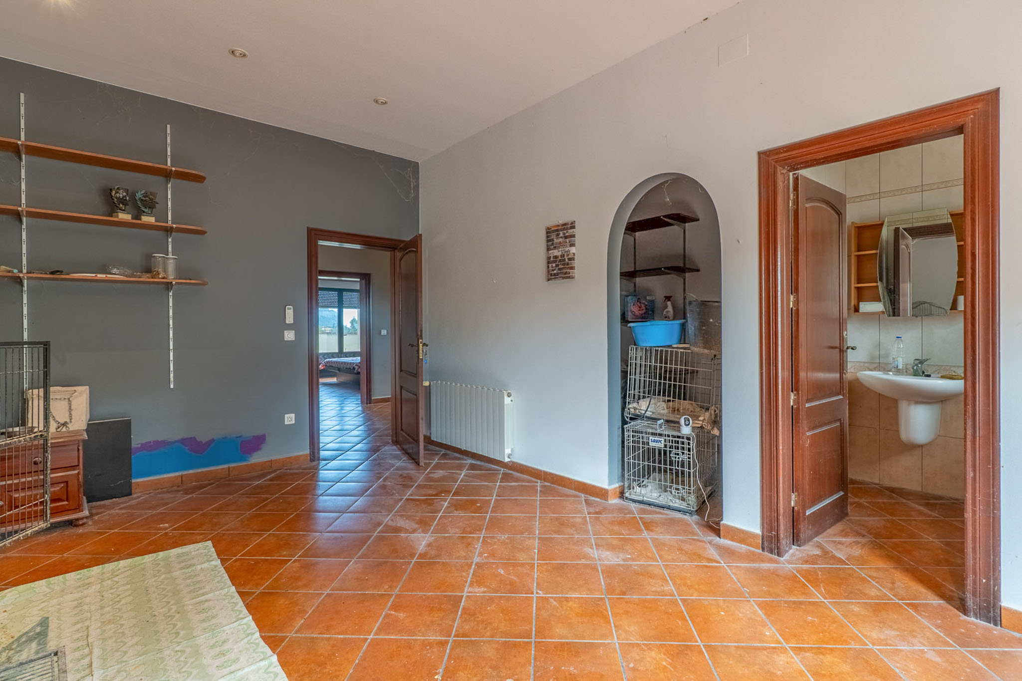 Interior room featuring terracotta floor tiles, two-tone walls, and wooden doors. It includes an arched alcove with shelves and provides access to a bathroom and another room.