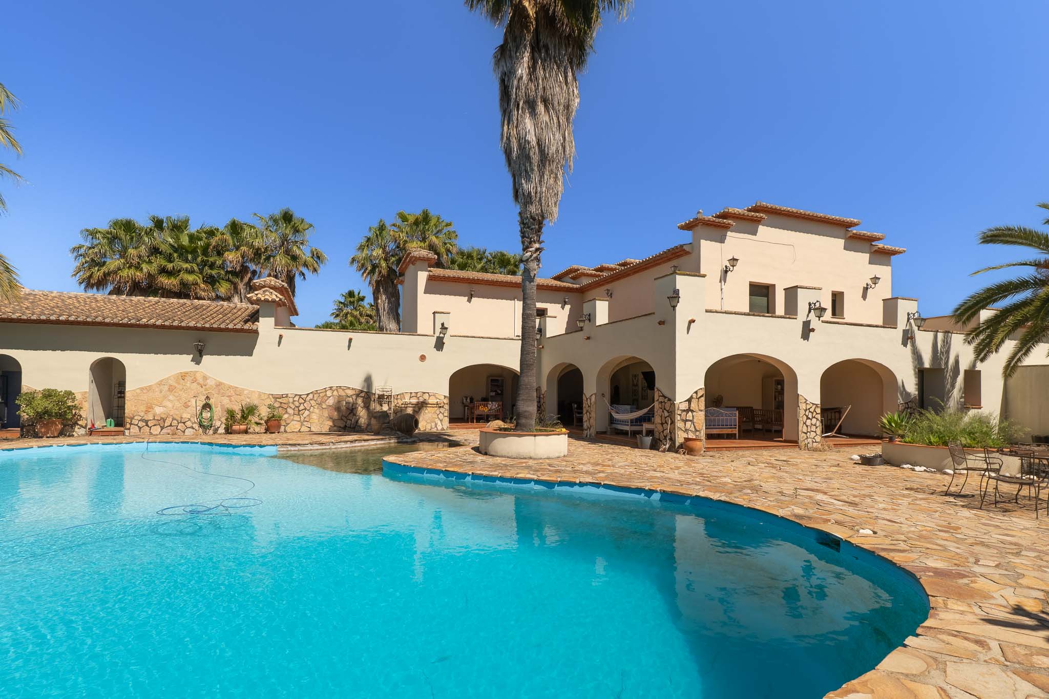 Irregularly shaped private swimming pool with clear blue water, bordered by an expansive natural stone terrace. The Mediterranean-style villa features arched loggias and terracotta roof tiles, amidst mature palm trees.