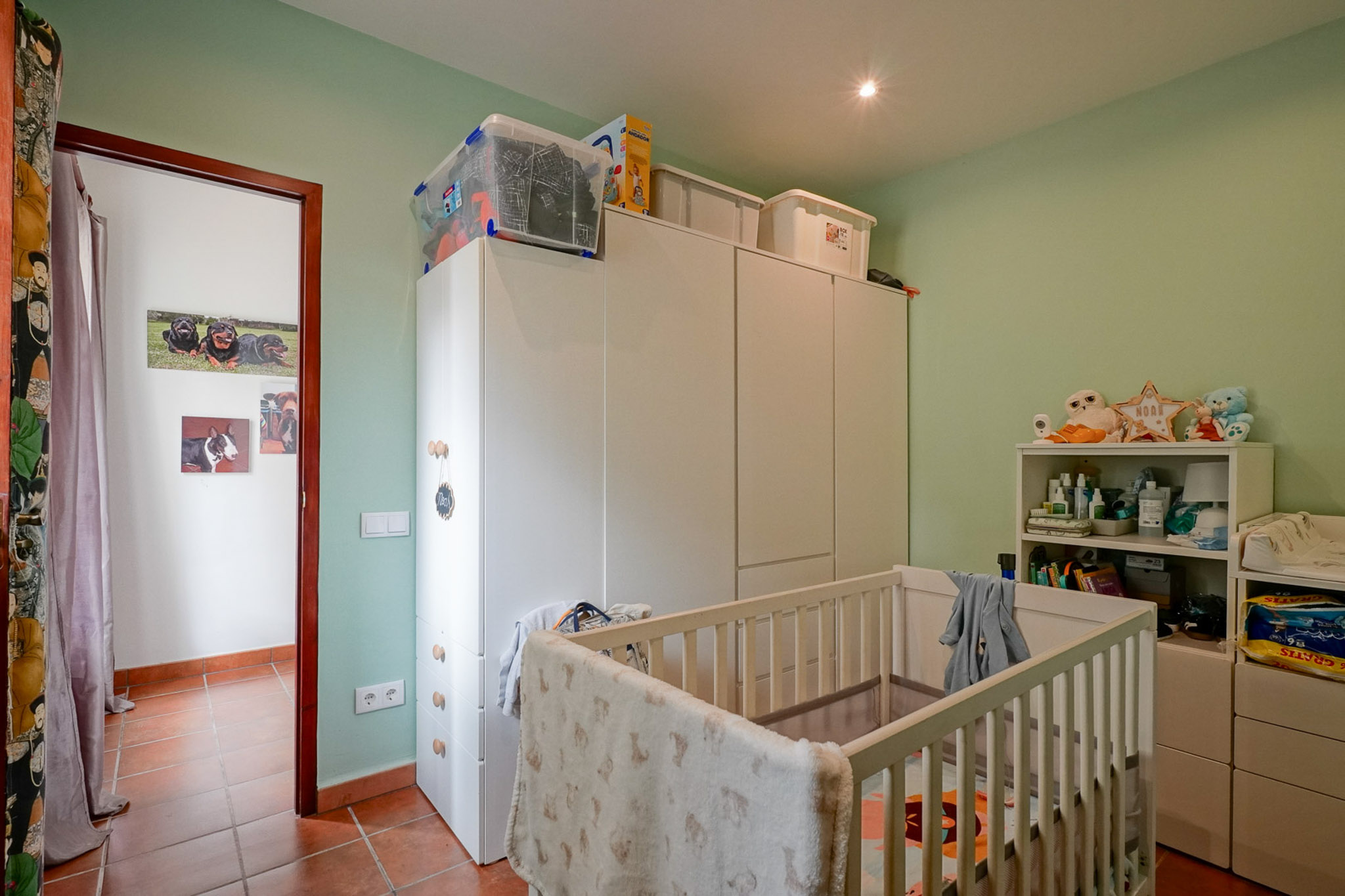 Nursery with light green walls, terracotta tiled floor, and a white crib. Features spacious white wardrobes, a shelving unit, and a changing table, illuminated by a recessed ceiling light.
