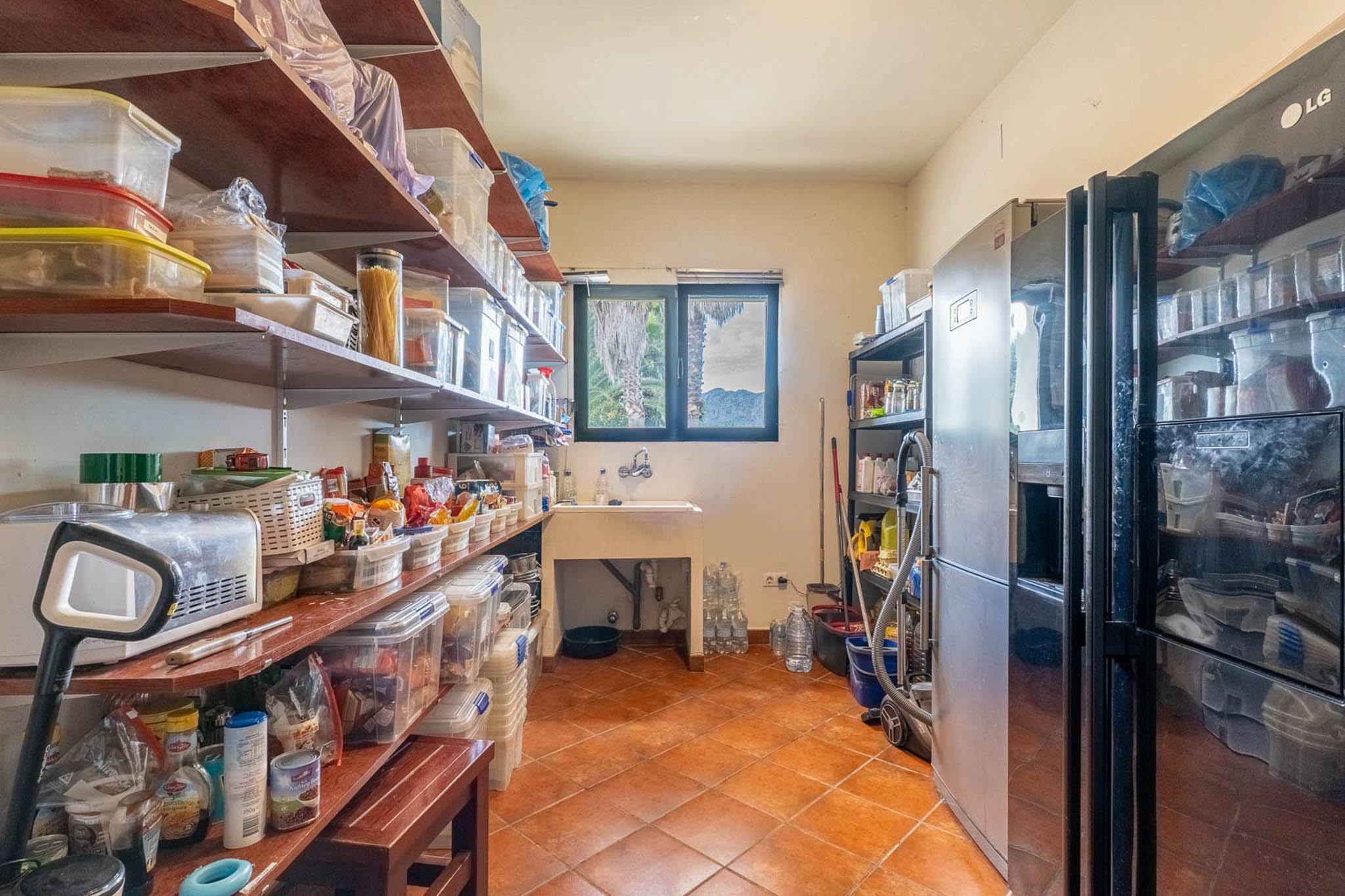 Practical pantry with extensive shelving, a utility sink, and two large refrigerators. Features terracotta tiled floor and a window with palm tree and mountain views.