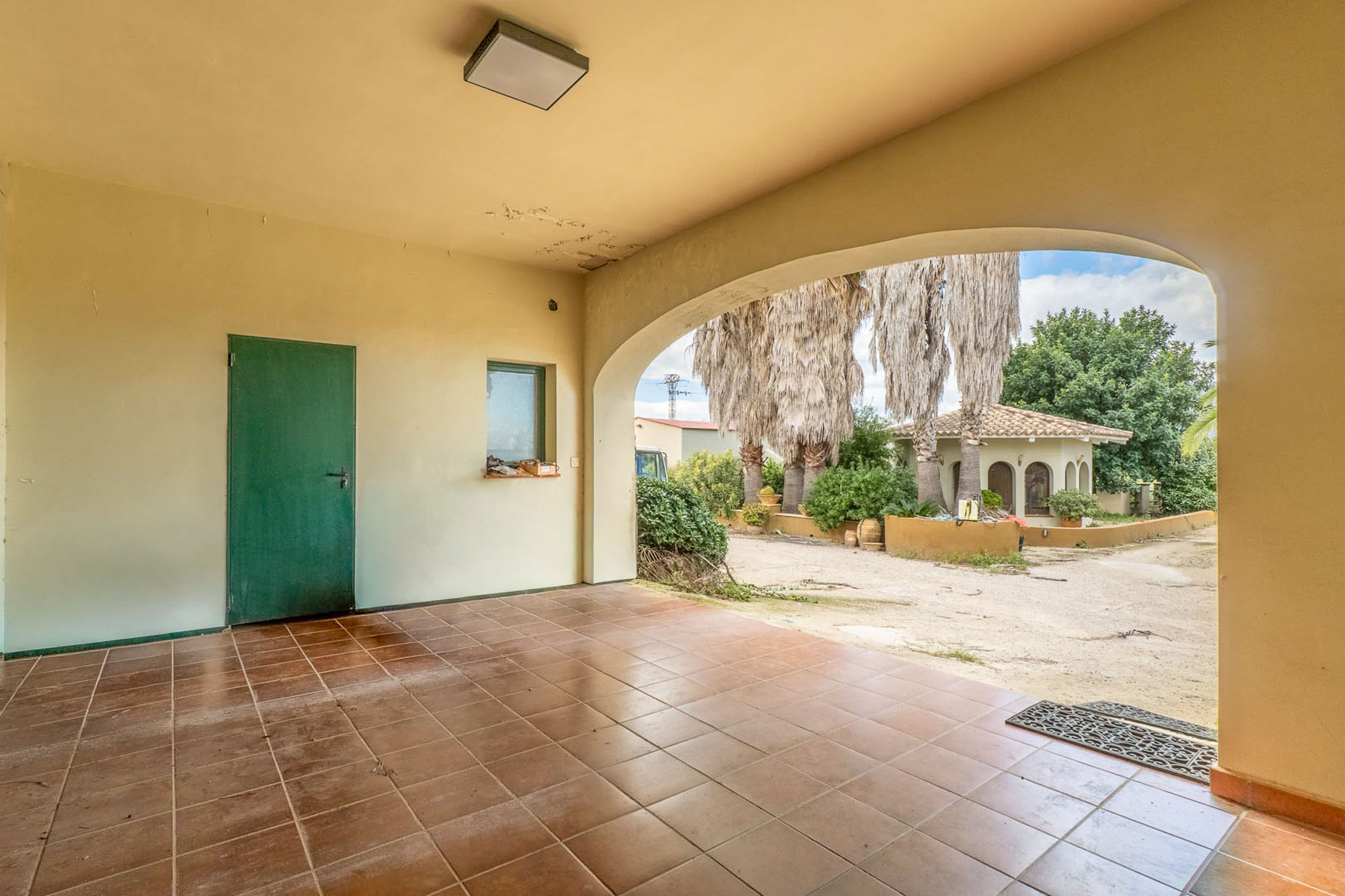 Spacious covered terrace featuring a tiled floor and an archway opening to a gravel path, palm trees, and an exterior building with a tiled roof.