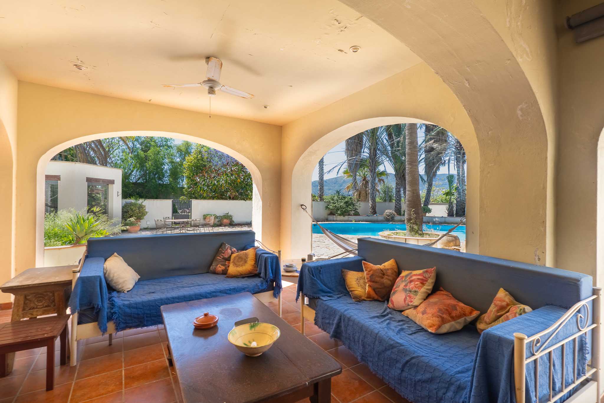 Spacious covered terrace with arches, terracotta flooring, and blue seating, offering views of the pool, palm trees, and distant hills.