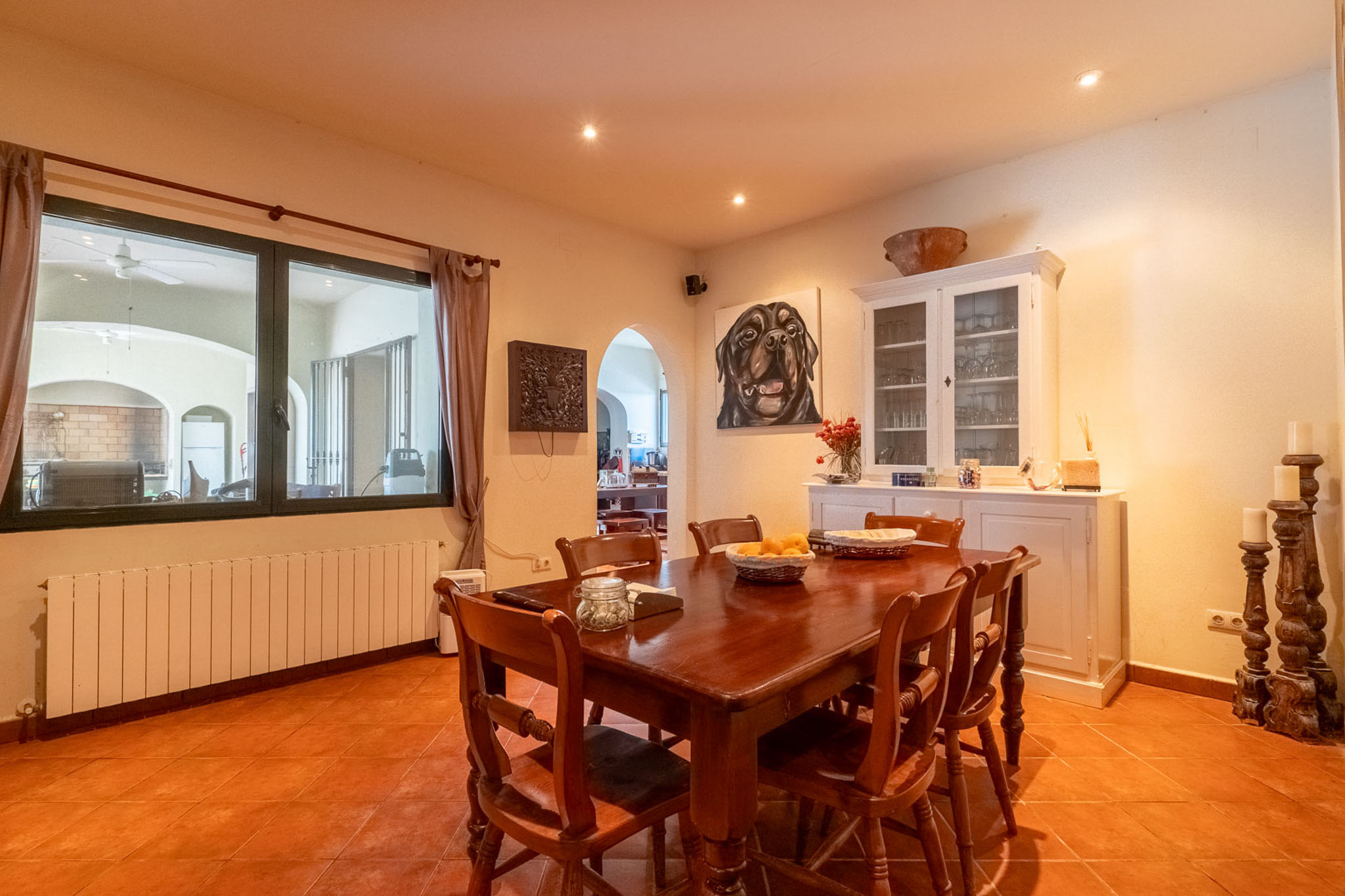 Spacious dining room featuring a dark wooden table and matching chairs, set on terracotta floor tiles. The area includes a radiator below the window and a white display cabinet.