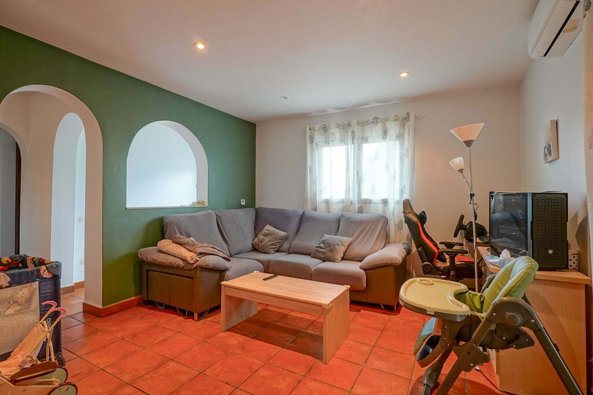 Spacious living area featuring a green accent wall with arched openings, terracotta tiled floor, and a comfortable corner sofa. Includes natural light and a dedicated entertainment zone.