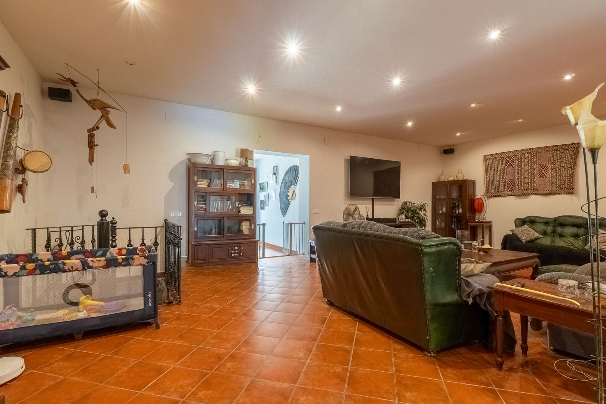 Spacious living area featuring terracotta tiled flooring, recessed ceiling lights, dark leather sofas, and wooden display cabinets. A wall-mounted television and an open archway to a staircase are visible.