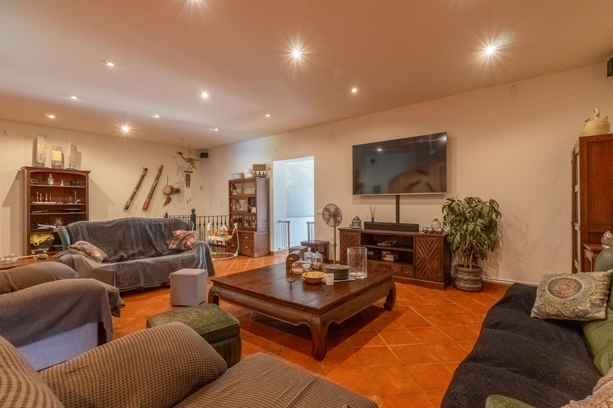Spacious living room featuring terracotta floor tiles, recessed ceiling spotlights, and a large wall-mounted TV. Furnished with dark wood pieces and covered sofas, creating a warm ambiance.