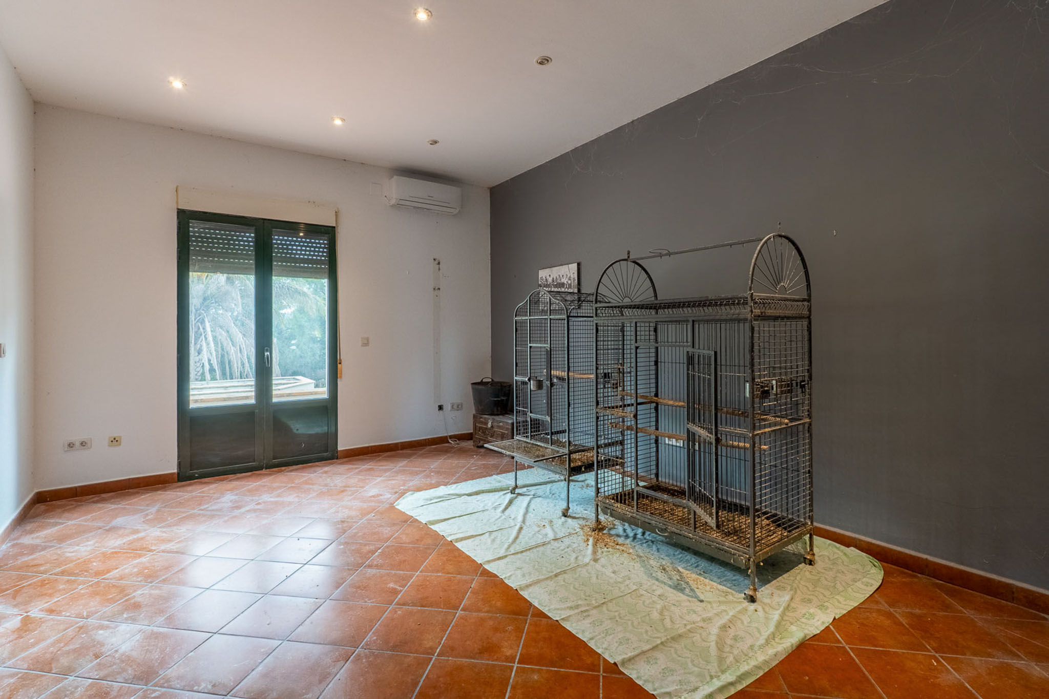 Spacious room with terracotta-tiled floor, two-tone walls, recessed lighting, and an air conditioning unit. Features a double-glazed door with roller shutter and large metal bird cages.