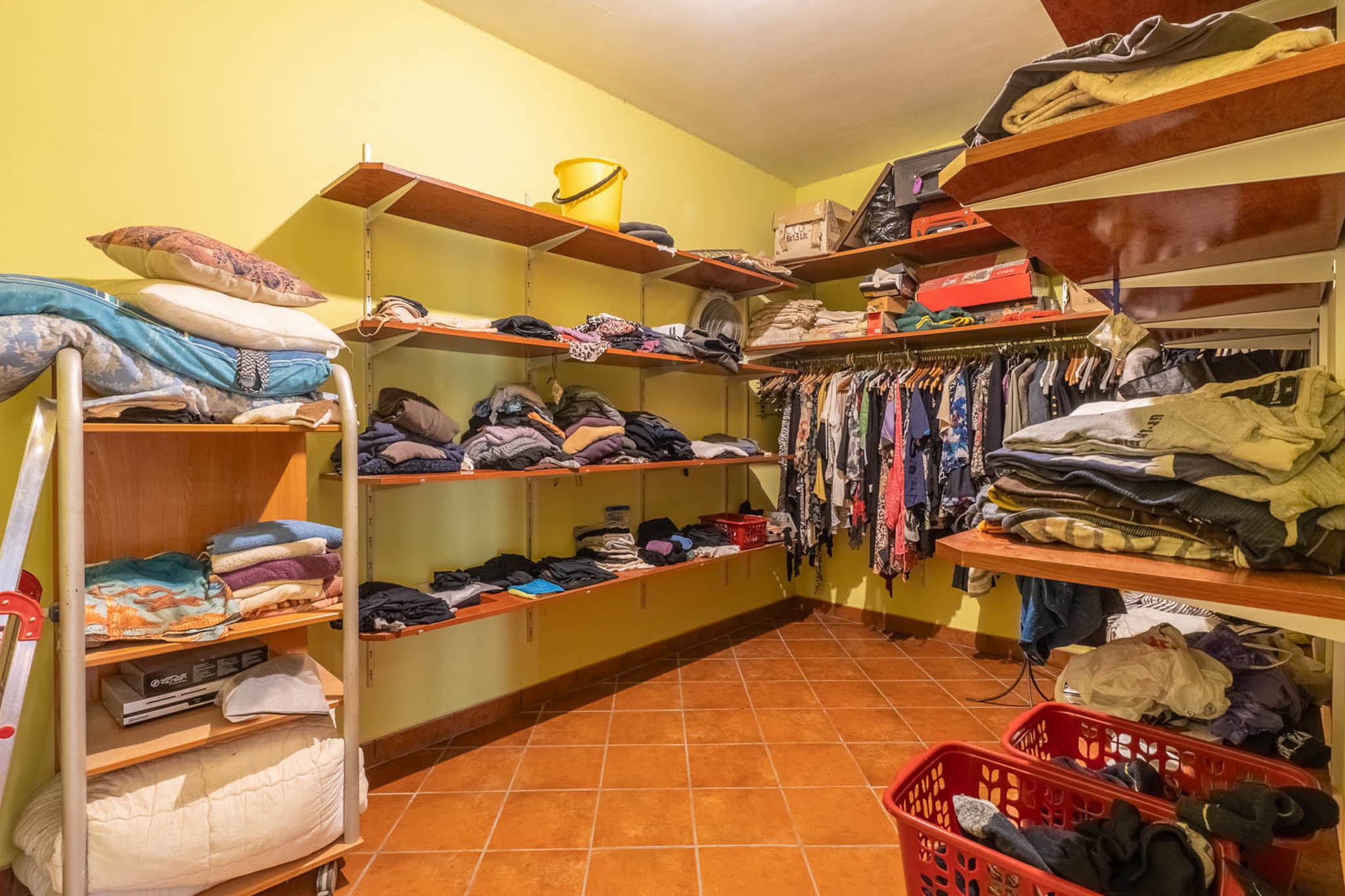 Spacious walk-in closet featuring open wooden shelving, light yellow walls, and terracotta floor tiles, providing extensive storage capacity.