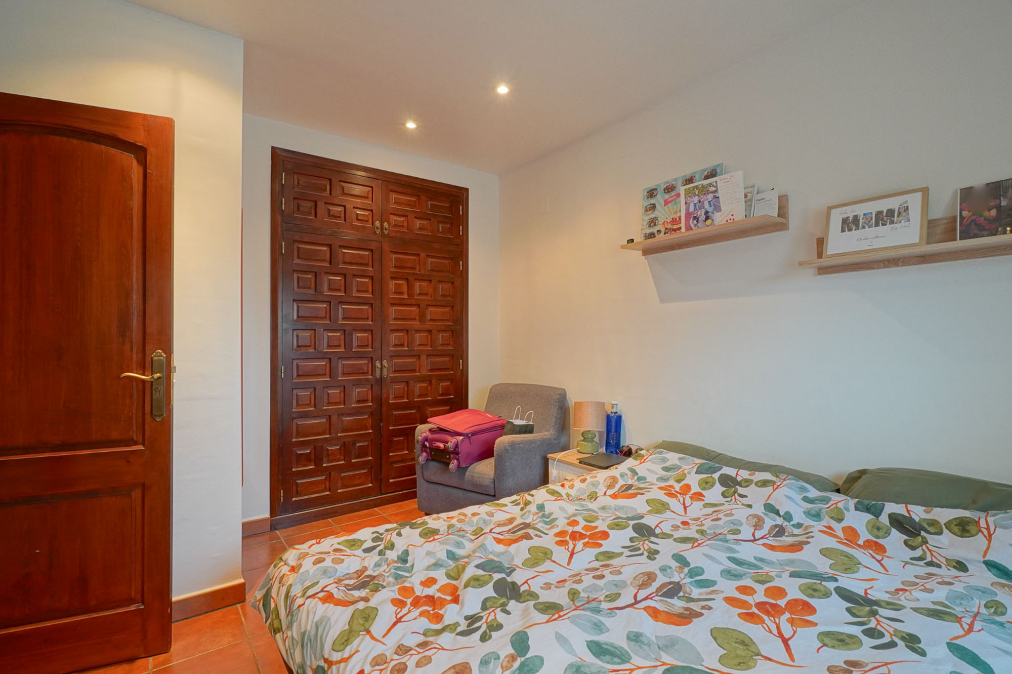 This bedroom features a dark paneled wooden wardrobe and door, terracotta floor tiles, and a bed with a vibrant leaf-patterned duvet. Recessed lighting illuminates the space.