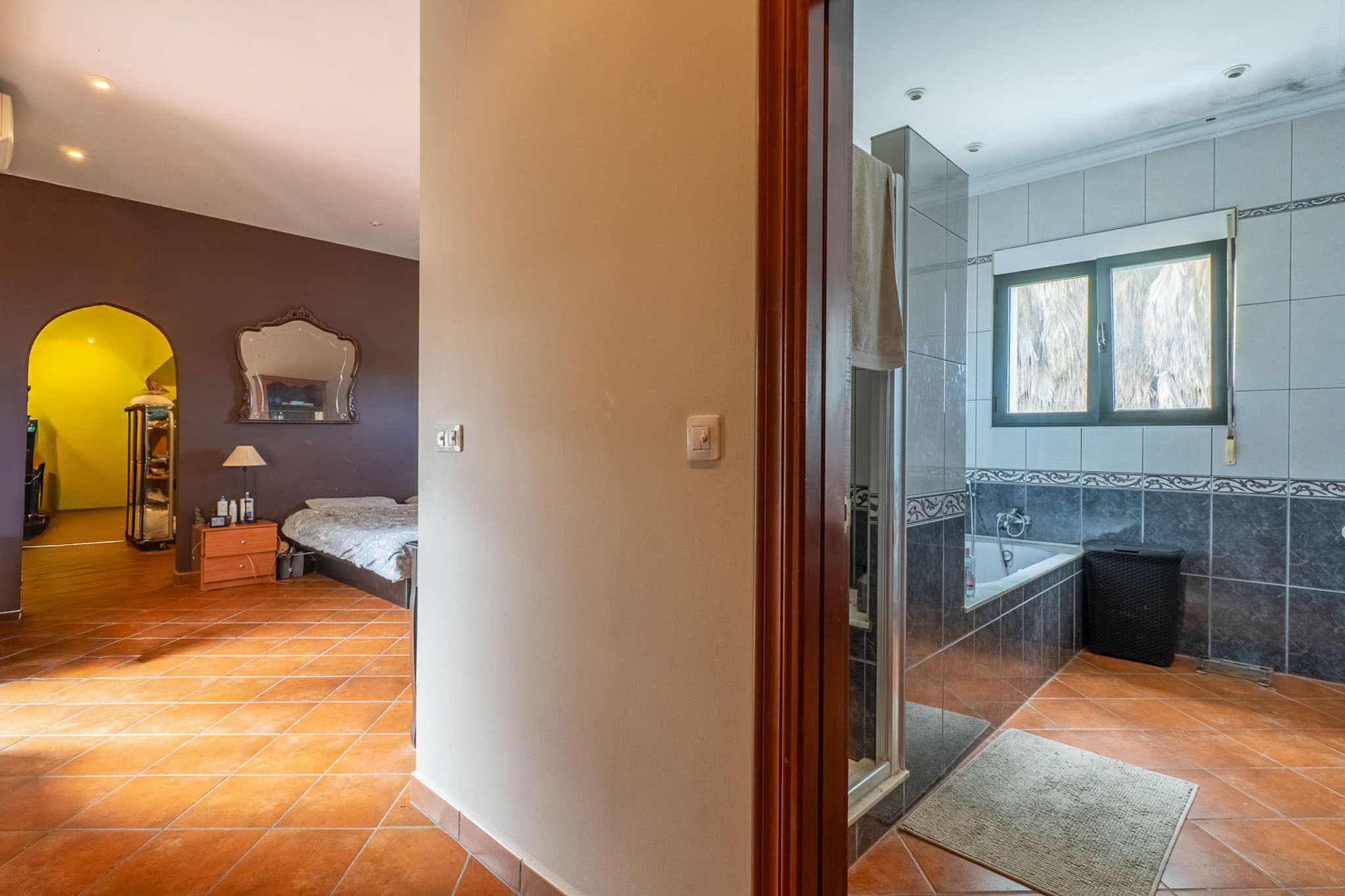 View from the hallway with terracotta tiles, showing a bedroom with dark walls and an ornate mirror on the left, and a tiled bathroom with a bathtub on the right.