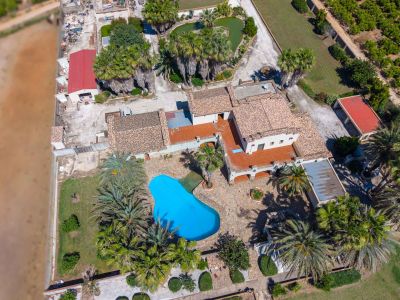 Aerial view of the estate featuring an irregular-shaped blue swimming pool, surrounded by palm trees and gardens. Multiple buildings with terracotta roofs and a heart-shaped pond are visible.