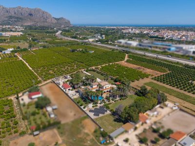 Aerial view of the estate featuring the main house, swimming pool, extensive gardens, and a large solar panel array, set amidst orange groves with distant sea and mountain views.